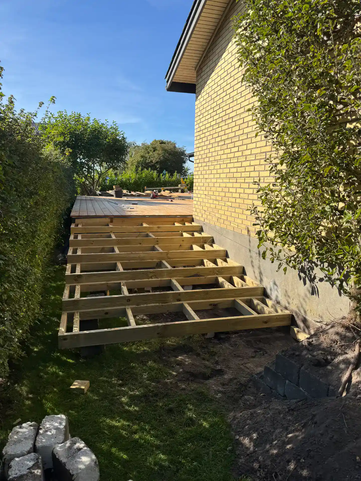 Træterrasse under opbygning ved husvæg med grønne omgivelser.