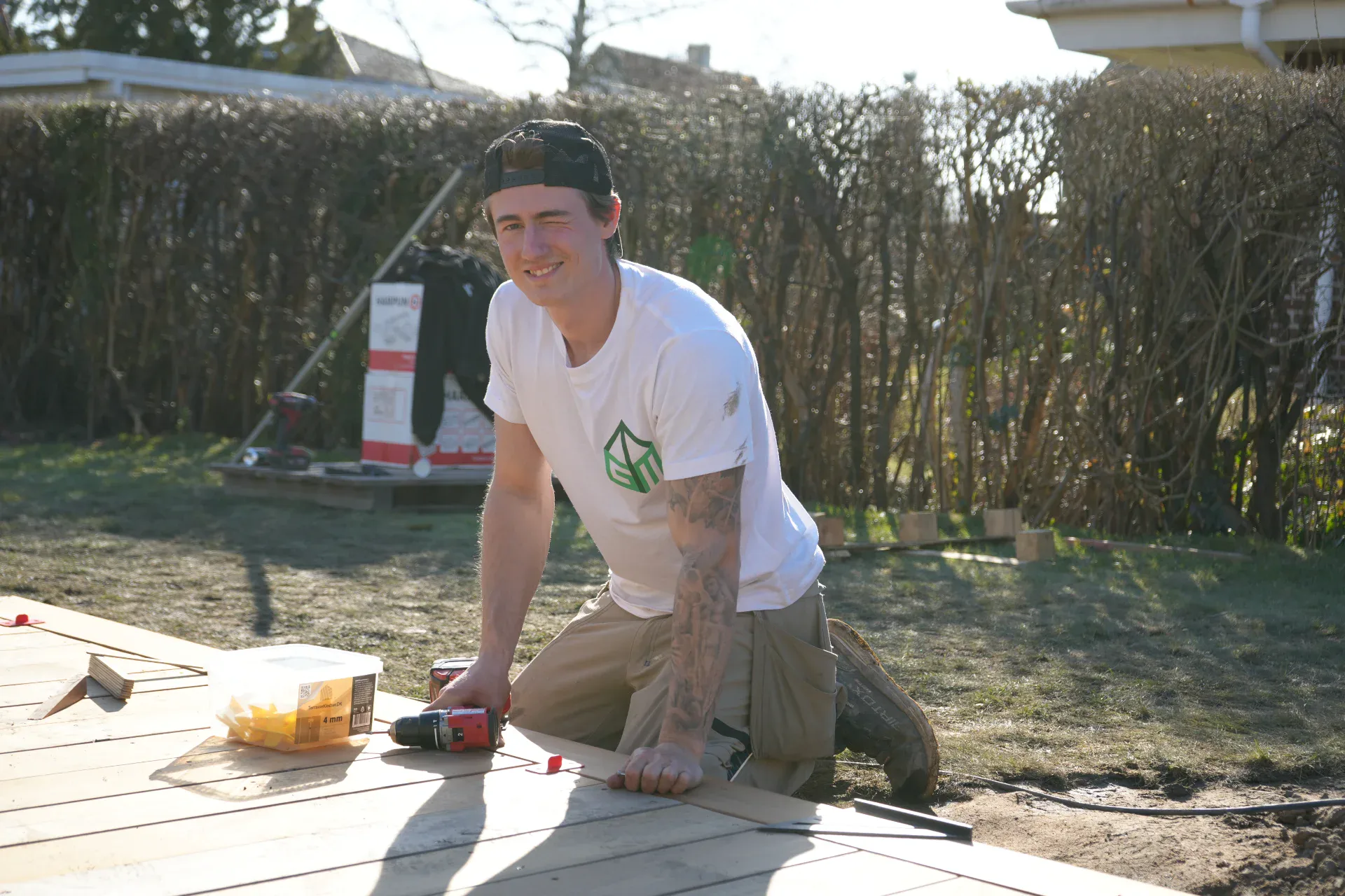 Håndværker i arbejdstøj, der arbejder på en træterrasse udendørs.